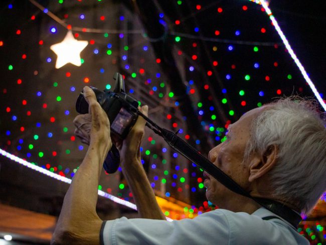 Ông Chuyên (80 tuổi, giáo xứ Nam Hòa) chăm chú ghi lại khung cảnh trong hẻm bằng chiếc máy ảnh kỹ thuật số.  "Giờ yếu rồi nên tôi chỉ đi gần nhà và cầm theo chiếc máy ảnh nhẹ nhất để đi chụp, đăng Facebook chơi. Đêm Giáng sinh, tôi mới thuê xe ôm chở đi xem vài con đường trang trí đẹp trong thành phố", ông nói.